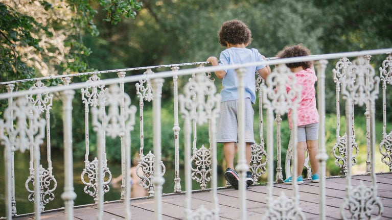Two children stand on the white bridge over the River Wandle at Morden Hall Park, London. Their backs can be seen as the look over the bridge railings to look in the water below.